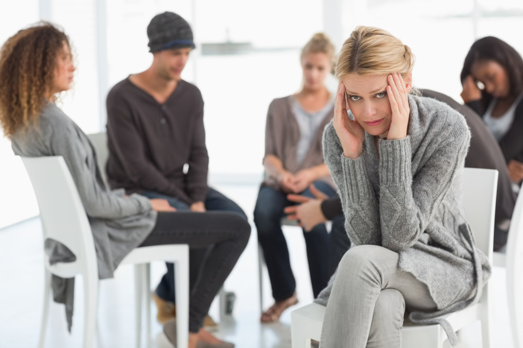 Upset woman with head in hands at rehab group looking at camera at therapy session